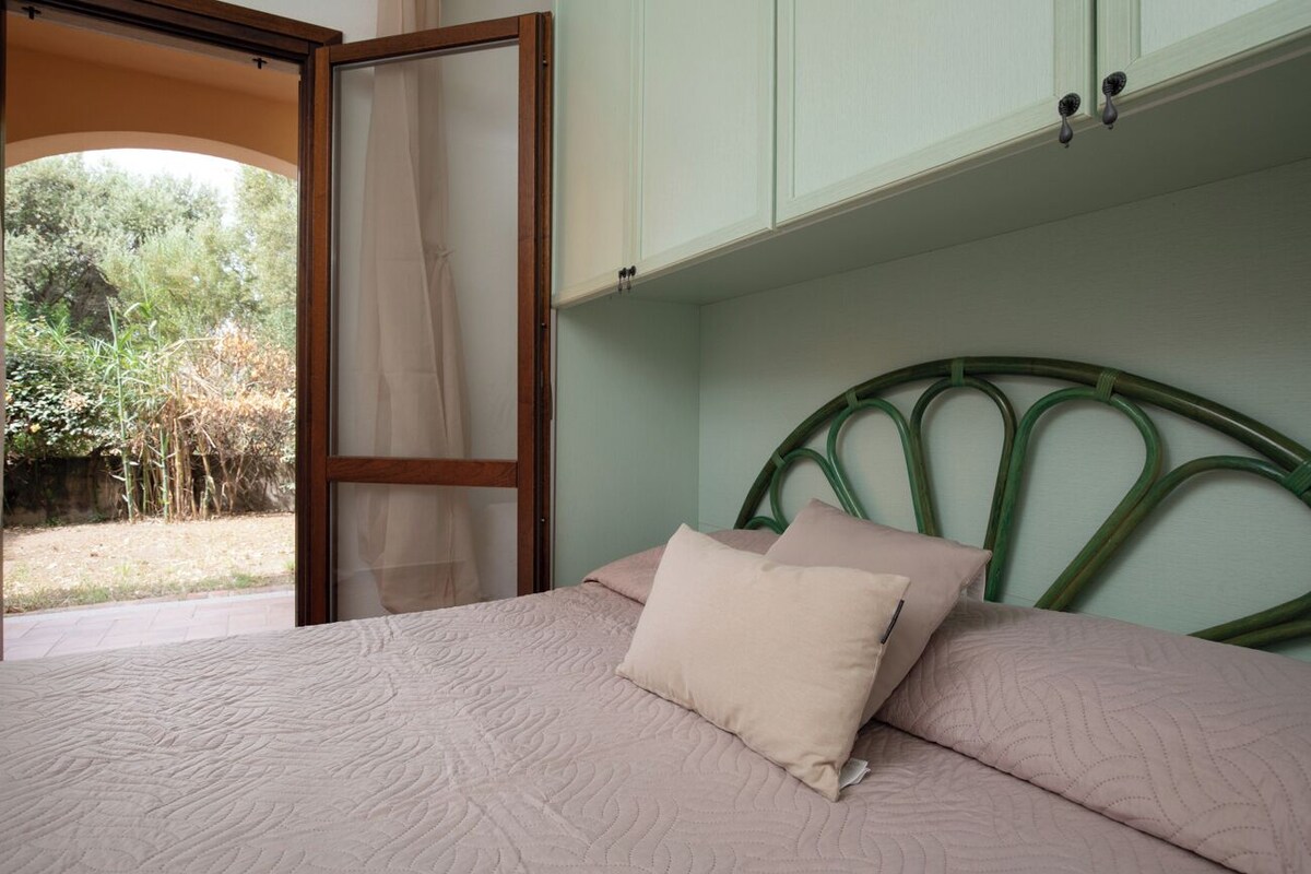A bed with a green metal headboard, a beige pillow, and a view of greenery outside.