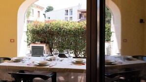 A dining table set with plates, glasses, and cutlery, with a view of buildings and a hedge through an archway.