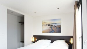 Een hotelkamer met een bed, twee bedlampen, een schilderij van een strandgezicht en een deur naar de badkamer.