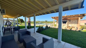 A covered patio with wicker furniture, a pool, and a view of a building with a stone facade. - Villa Aurelia | Private Pool, 10 Guests, Near the Sea (Sicilia)
