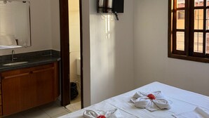 Ein Hotelzimmer mit Bett, Deckenventilator, Wand-Fernseher, Fenster und einem Badezimmer mit Waschbecken und Spiegel.