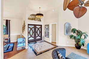 A spacious living area with a large wooden door, a ceiling fan, a chandelier, and a potted plant.