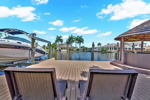 A wooden deck with two wicker chairs, a boat docked on the left, and a tiki bar on the right. - VRCC Villa Waterfall (Cape Coral)
