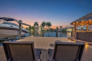 Una terraza de madera con dos sillones de mimbre, un puerto deportivo con barcos y vistas al atardecer.