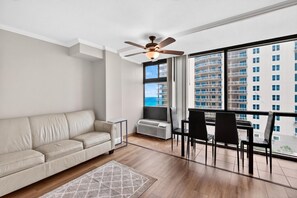 A modern living room with a beige sofa, a ceiling fan, a dining table with chairs, and large windows offering a view of a high-rise building. - 🌴Amazing Ocean Views | Beachfront Pool | Oceanside Inn 606🌴 (Daytona Beach)