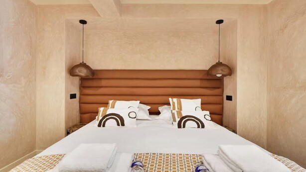 Ein Hotelzimmer mit einem Holz-Kopfteil, zwei Pendelleuchten, einem ordentlich bezogenen Bett mit gemusterten Bettwäsche und einem kleinen Tisch mit Handtüchern und einer Flasche.