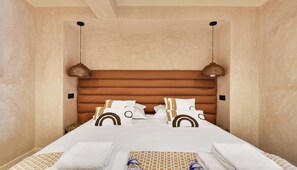 A hotel room with a wooden headboard, two pendant lights, a neatly made bed with patterned bedding, and a small table with towels and a bottle. - COMBINING MODERN COMFORT WITH AN EXCEPTIONAL CENTRAL LOCATION -  RUE DU CAIRE (Paris)