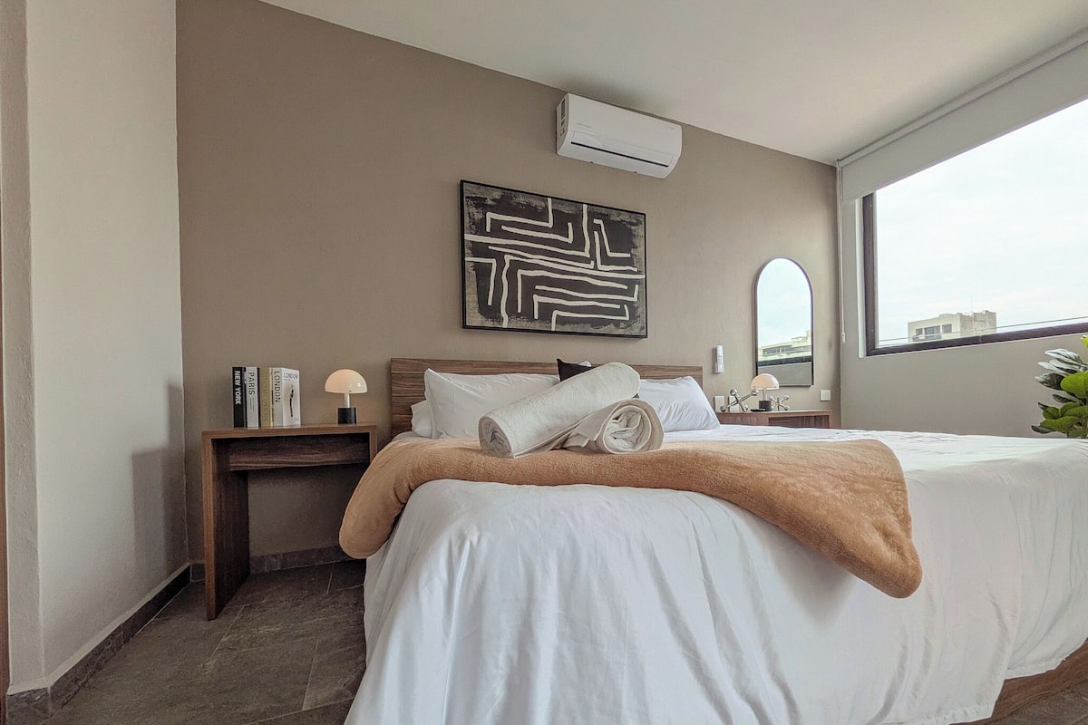 Une chambre d’hôtel avec un grand lit, une table de chevet avec une lampe, un climatiseur fixé au mur et une grande fenêtre.