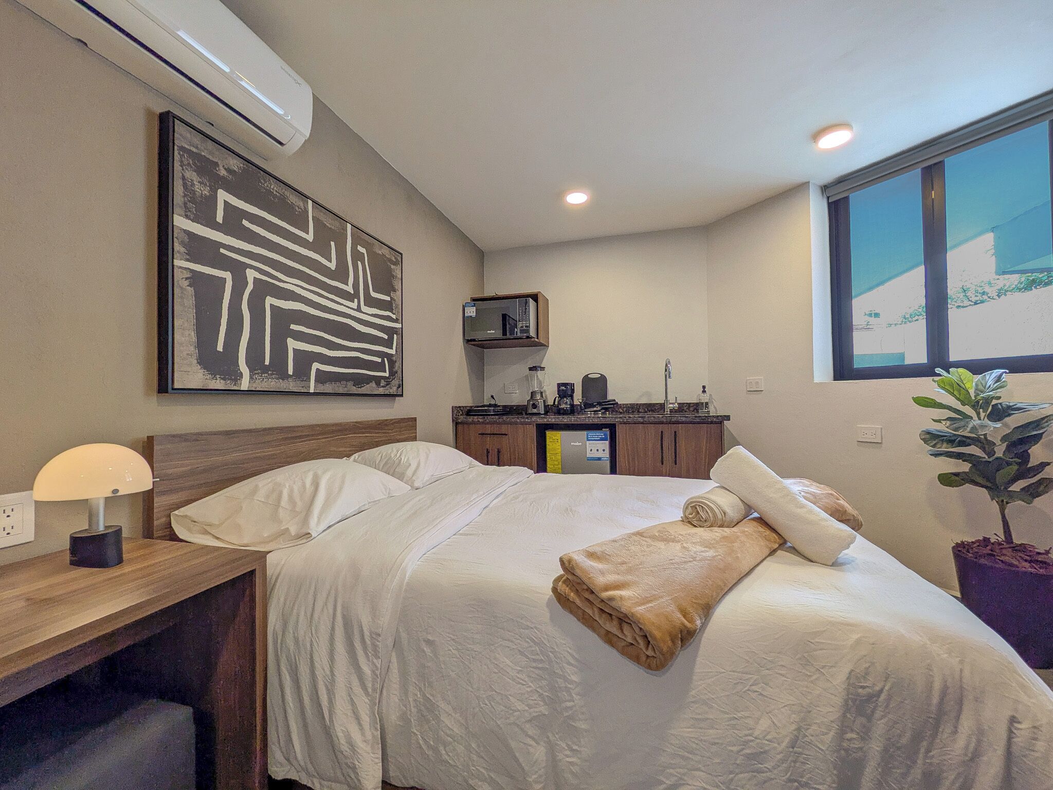 A hotel room with a bed, bedside table, lamp, air conditioning unit, kitchenette, and a window with a view.