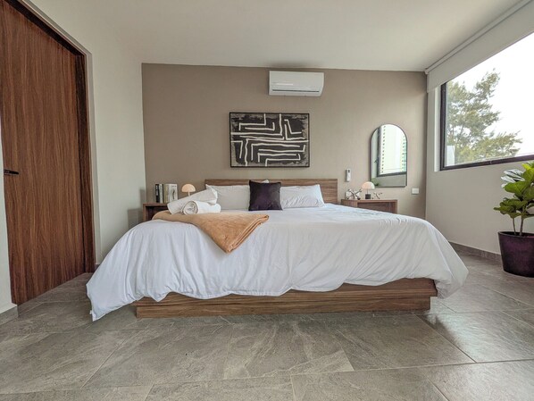 Une chambre moderne avec un grand lit, une tête de lit en bois, un climatiseur fixé au mur, une table de chevet avec une lampe et une fenêtre donnant sur des arbres.