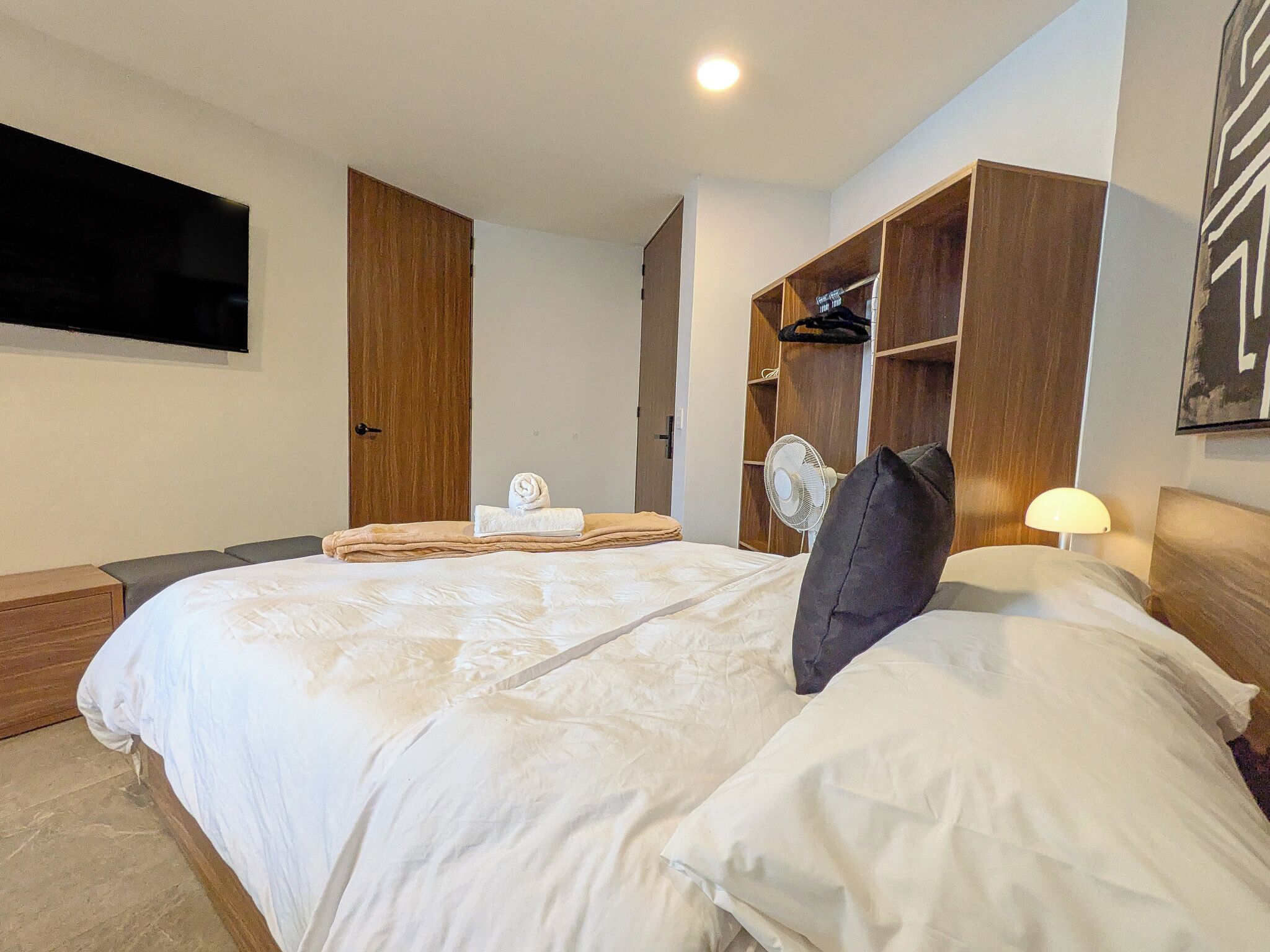 Quarto de hotel com uma cama grande, um guarda-roupa, uma TV fixada na parede e um ventilador.