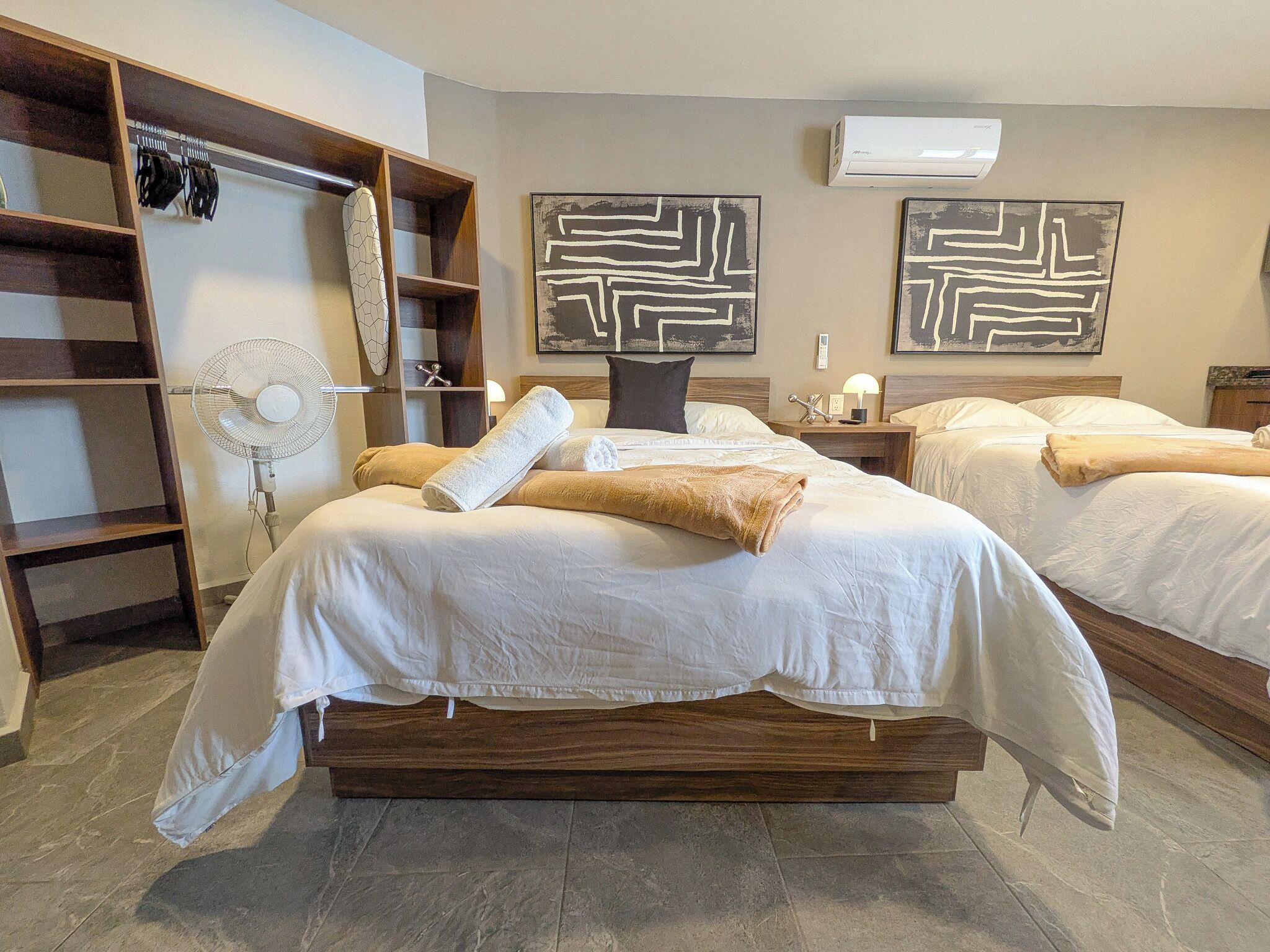 A hotel room with a large bed, a bedside table, a lamp, a fan, and abstract wall art.