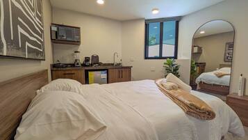 A bedroom with a bed, a bathroom, a kitchenette, and a large window.
