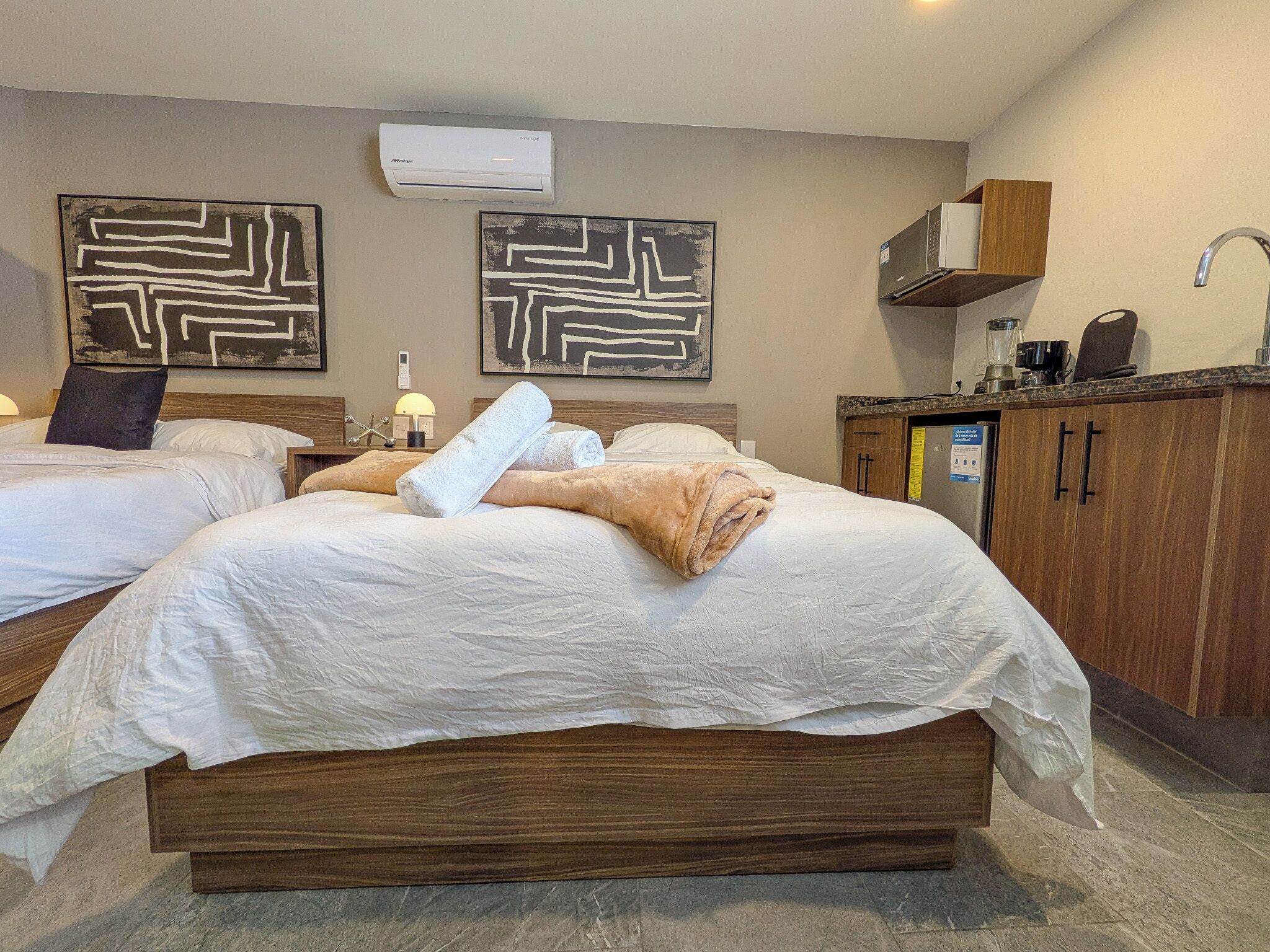 Quarto de hotel com duas camas, cabeceira de madeira, secretária embutida, cozinha compacta e ar-condicionado fixado na parede.
