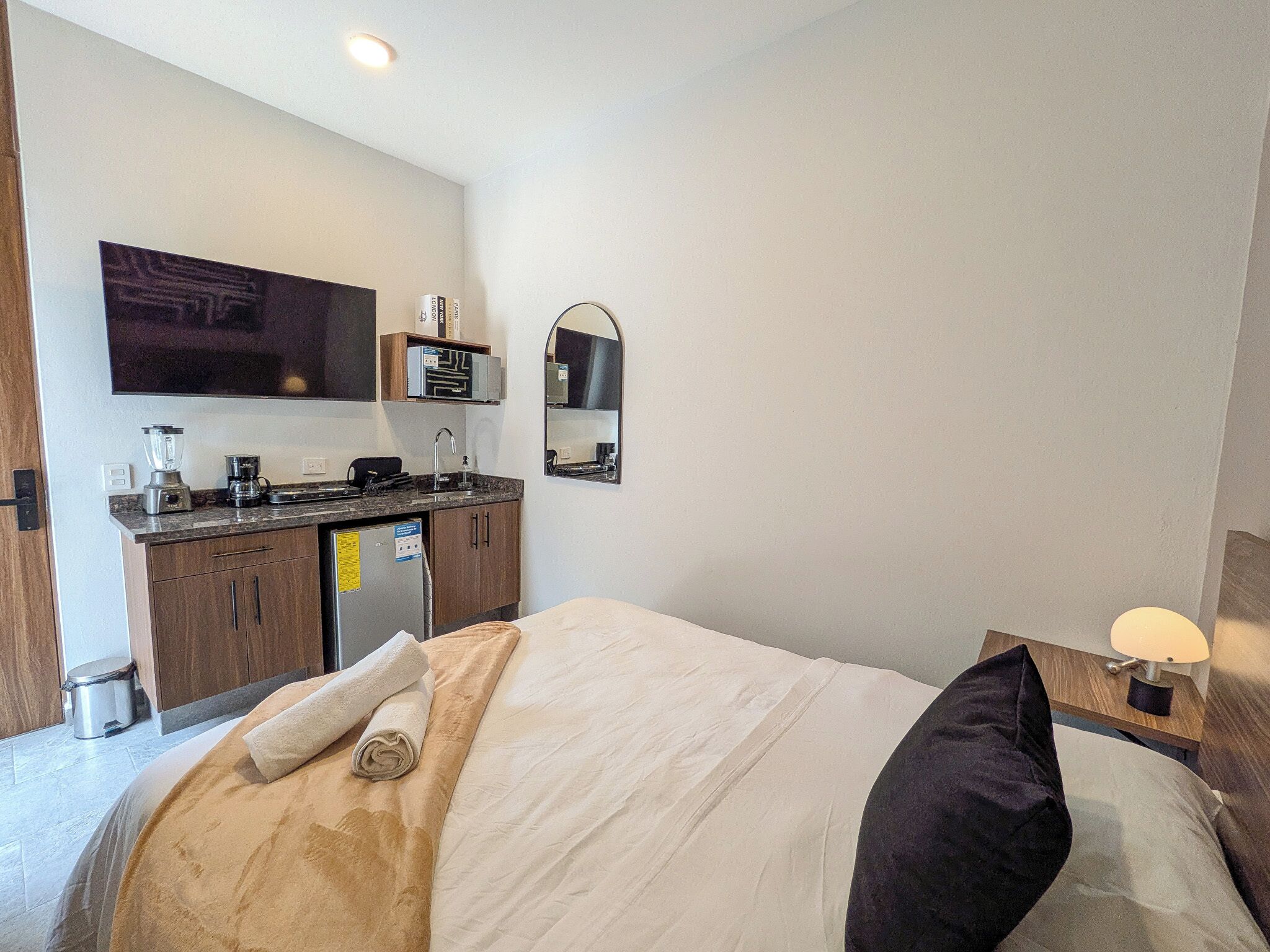 Une chambre d’hôtel comprenant un lit, une petite kitchenette, un miroir et une table de chevet avec une lampe.