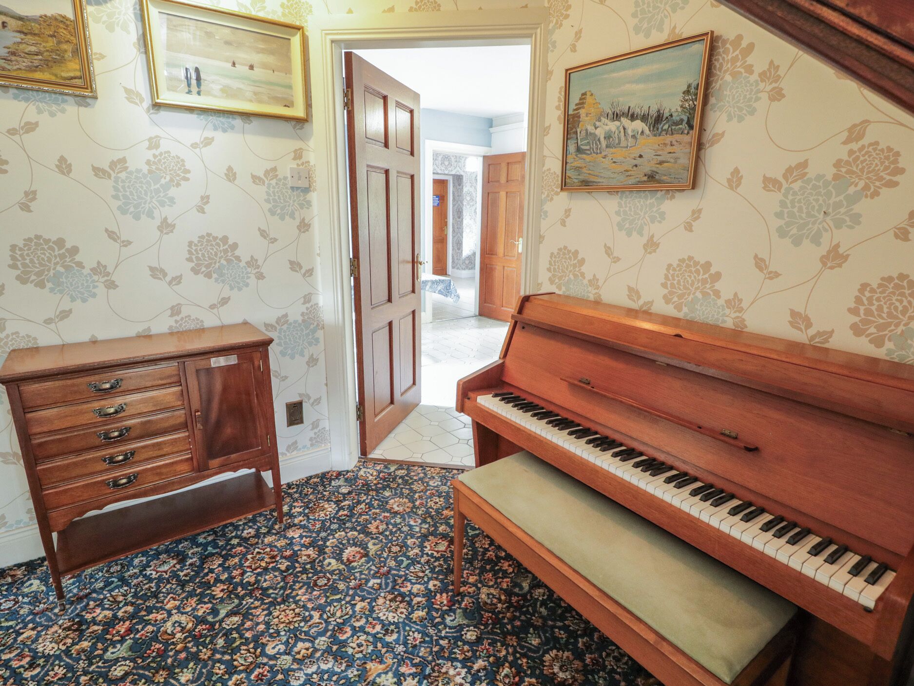 A room with a wooden piano, a cabinet with drawers, floral wallpaper, and a patterned carpet.