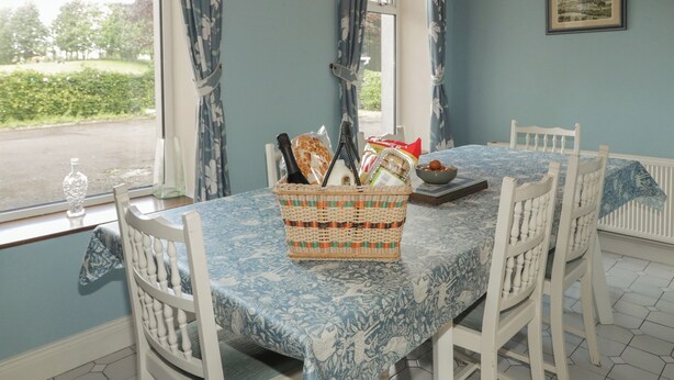 Un coin repas avec une table dressée pour quatre personnes, un panier en osier contenant des aliments, une horloge au mur et une fenêtre avec des rideaux.