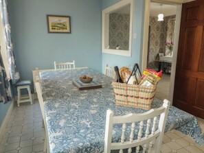 A dining area with a table set for four, a wicker basket with food items, a framed landscape picture, and a white chair.