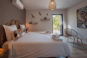 A bedroom with a large bed, a wicker chair, a small table, and a view of greenery outside.