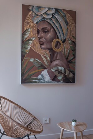 A wall-mounted painting of a woman with a headscarf and gold jewelry, a wicker chair, and a small wooden table with a plant.