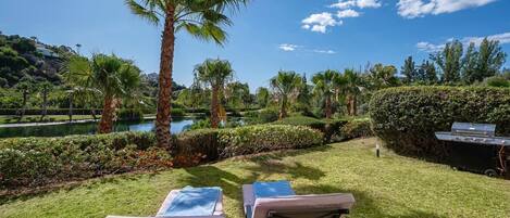 A patio with two cushioned chairs, a grill, and a view of a lake and palm trees.