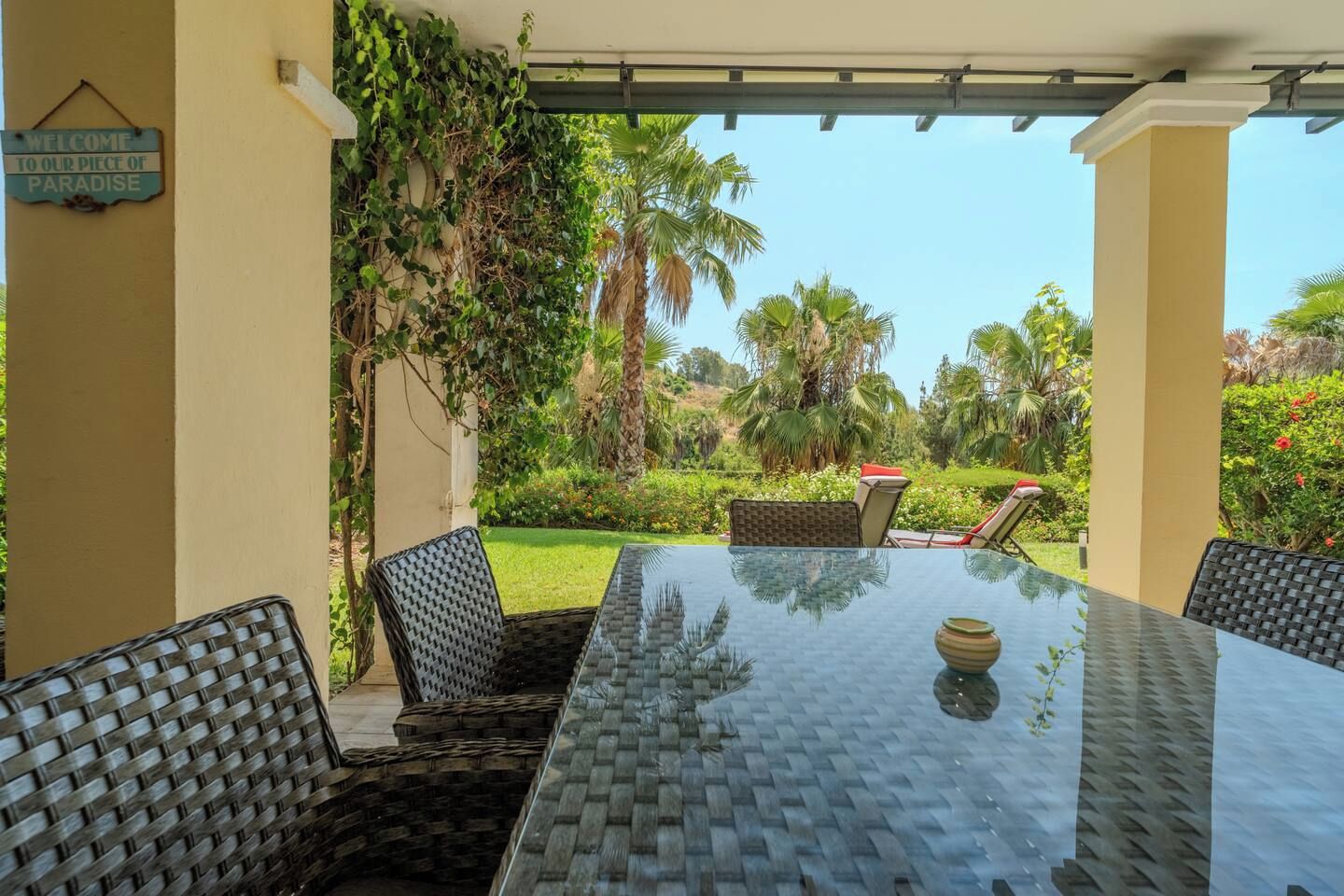 Eine Terrasse im Freien mit einem Glastisch und Korbstühlen, umgeben von üppiger Vegetation und Palmen.