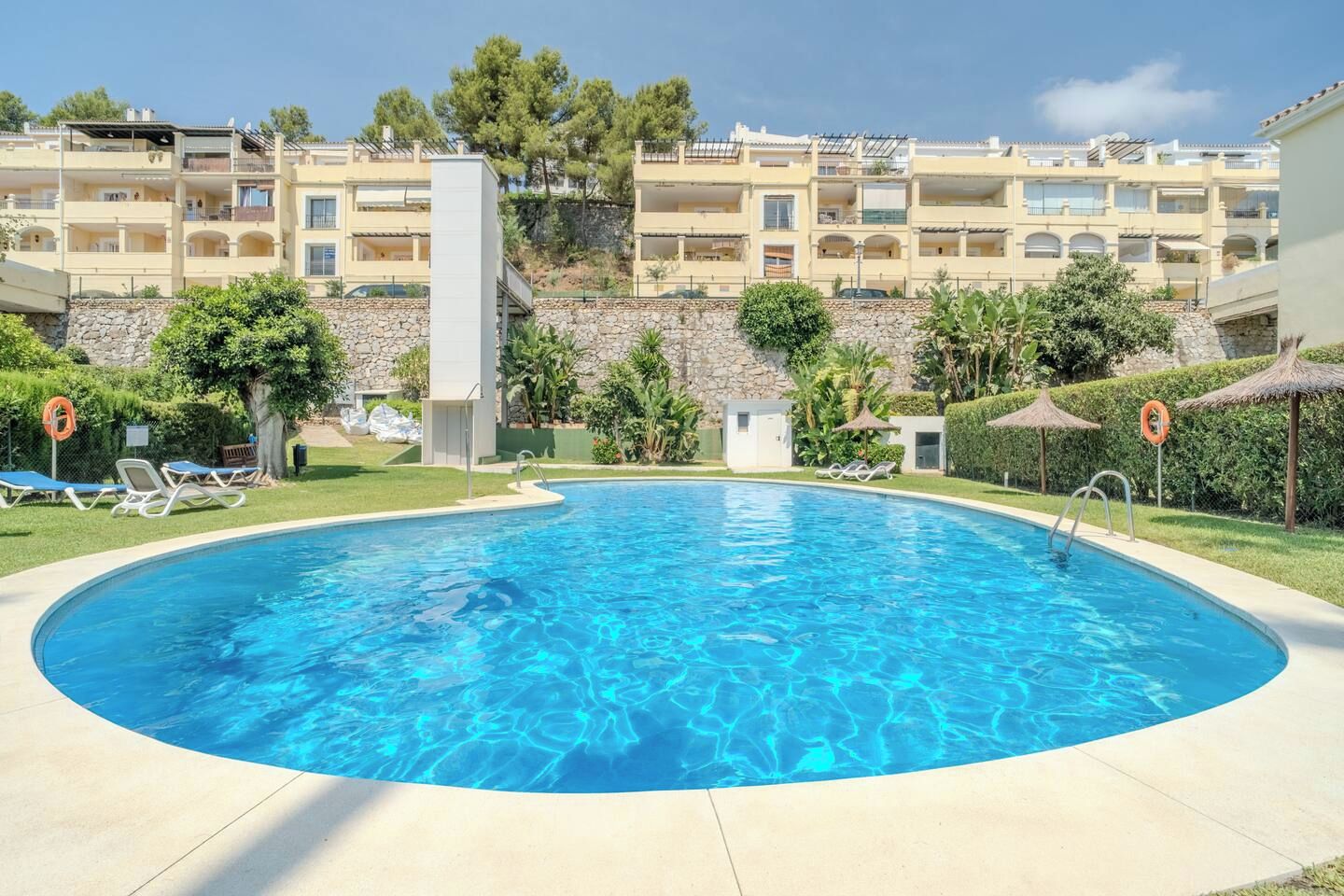 Una piscina con sillones de descanso y sombrillas, rodeada por un muro de piedra y vegetación.