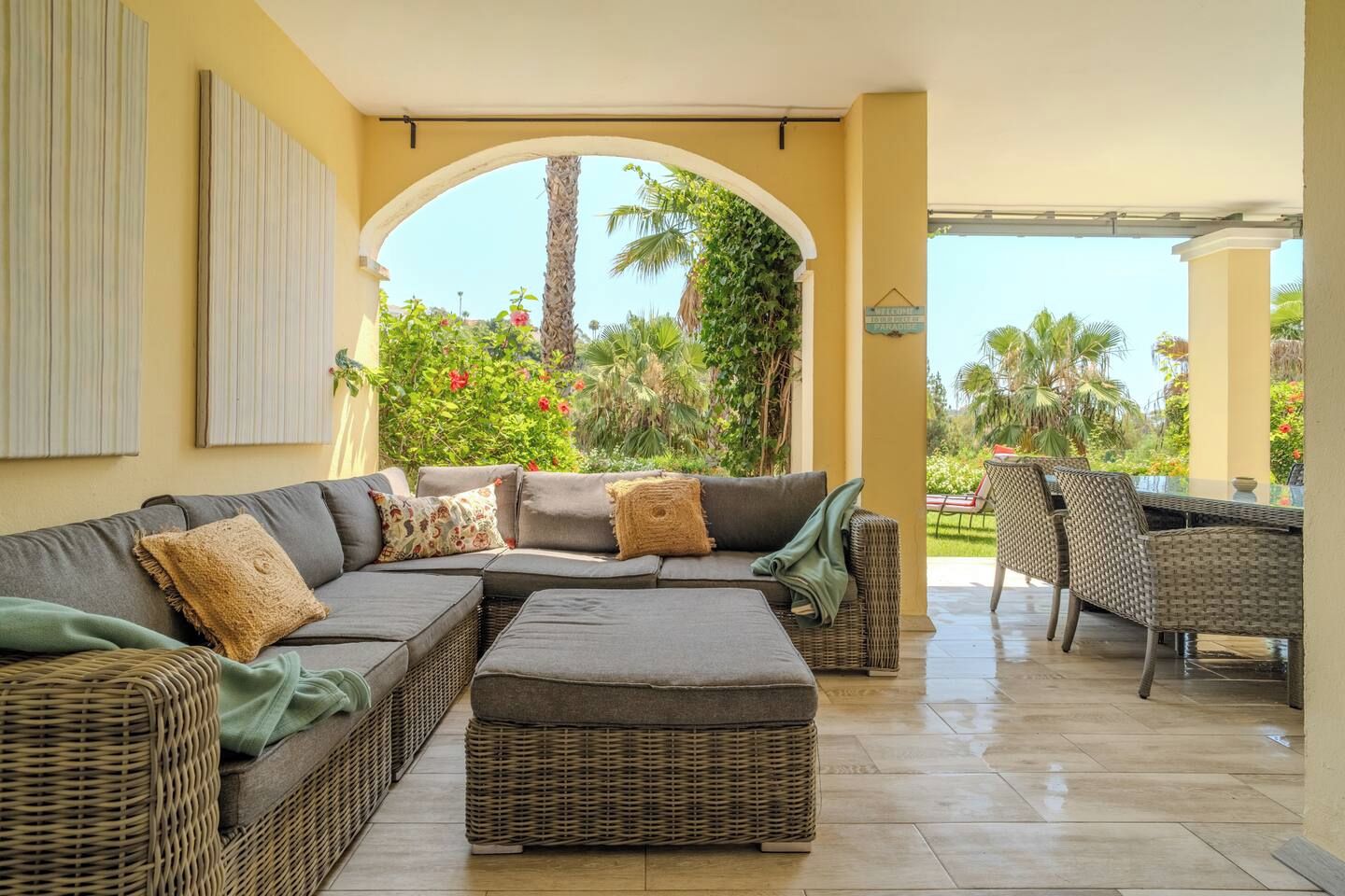 Un espacio de estar al aire libre techado, con un sofá y sillones, piso de baldosas y vistas a zonas verdes.