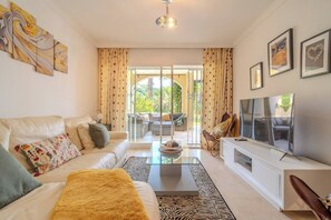 A modern living room with a sofa, coffee table, and a flat-screen TV. There is a glass door leading to an outdoor area with seating. - Lalq13 Stunning Frontline Golf 3 bed apt (Benahavis)