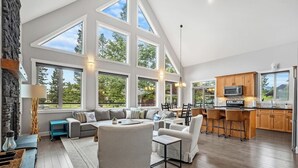 A spacious living area with a large window, a kitchen with wooden cabinets, and a seating area with a checkered ottoman. - Evercrest A-Frame | Hot Tub | Pool Tbl | Fireplace (Radium Hot Springs)