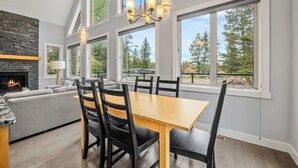 A modern interior with a fireplace, dining table, and large windows offering views of trees and a house. - Evercrest A-Frame | Hot Tub | Pool Tbl | Fireplace (Radium Hot Springs)