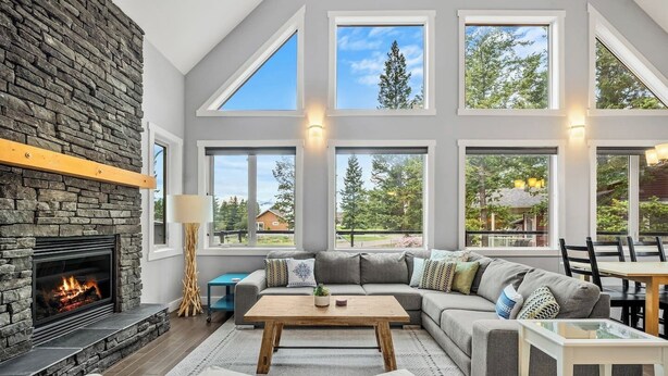 A modern living room with a stone fireplace, large windows, a sofa set, and a dining area. - Evercrest A-Frame | Hot Tub | Pool Tbl | Fireplace (Radium Hot Springs)