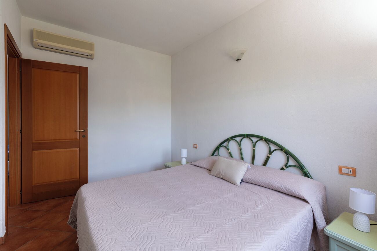 Un dormitorio con una cama, una puerta de madera, una mesita de noche con una lámpara y un aire acondicionado instalado en la pared.
