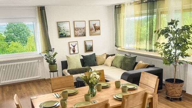 A dining area with a wooden table set for four, a sofa, and a large window with a view of greenery.