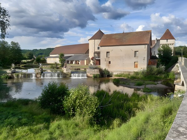A riverside building with a small waterfall, surrounded by greenery and a clear sky.