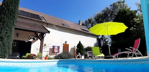 A swimming pool with lounge chairs and an umbrella, a white building with a brown roof, and trees in the background.