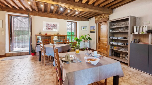 A dining area with hexagonal tile flooring, wooden tables, and chairs. There is a bookshelf with glassware and books, and a window with a view of greenery.