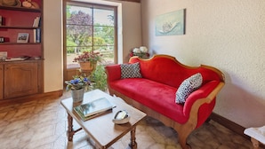 A cozy room with a red sofa, a wooden coffee table, a bookshelf, and a window with a view of greenery.