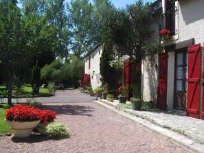 A pathway leading to a building with red shutters, surrounded by greenery and potted plants.