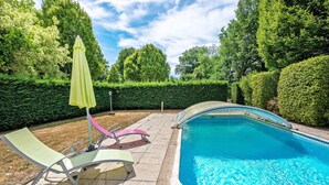 A swimming pool with a curved edge, surrounded by a paved patio with lounge chairs and a green umbrella.