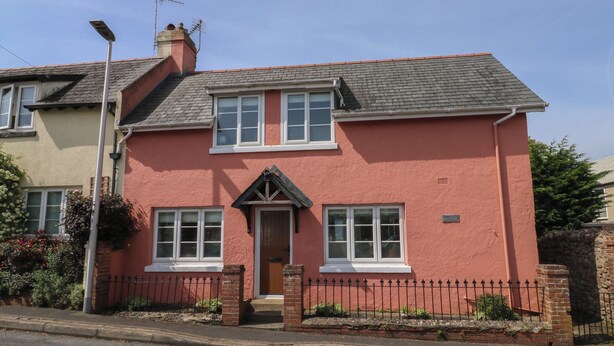 A two-story house with a red exterior, a front door, and a small porch. - Tracey Cottage (Newton Abbot)
