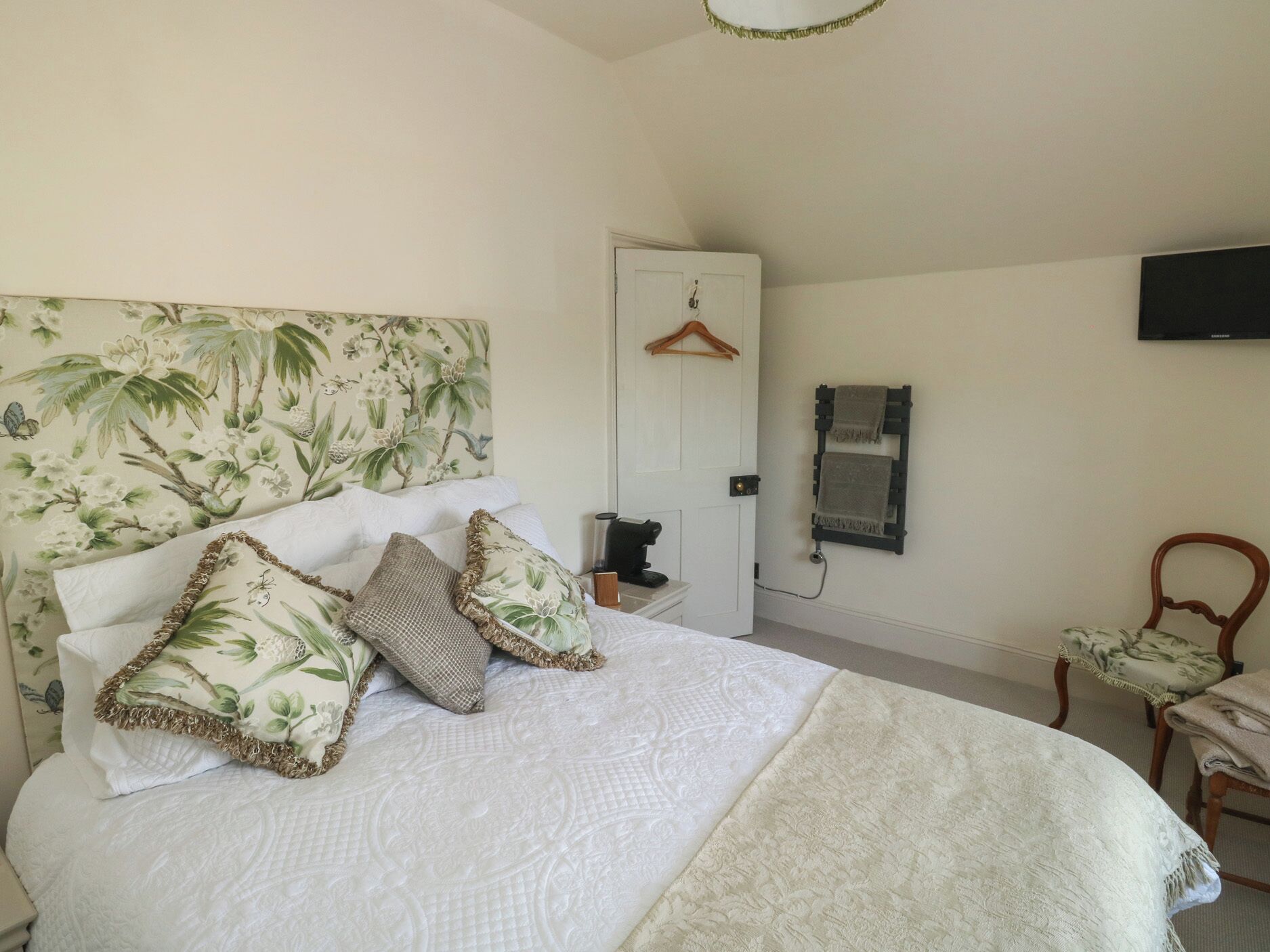 A bedroom with a bed, pillows, a bedside table, a chair, a coat rack, and a wall-mounted TV.