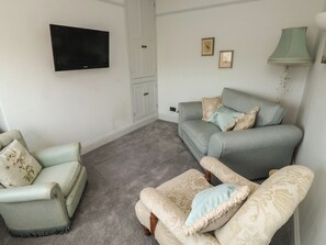 A living room with a grey sofa, a light green armchair, and a matching ottoman. A flat-screen TV is mounted on the wall, and there are two framed pictures on the wall. - Tracey Cottage (Newton Abbot)