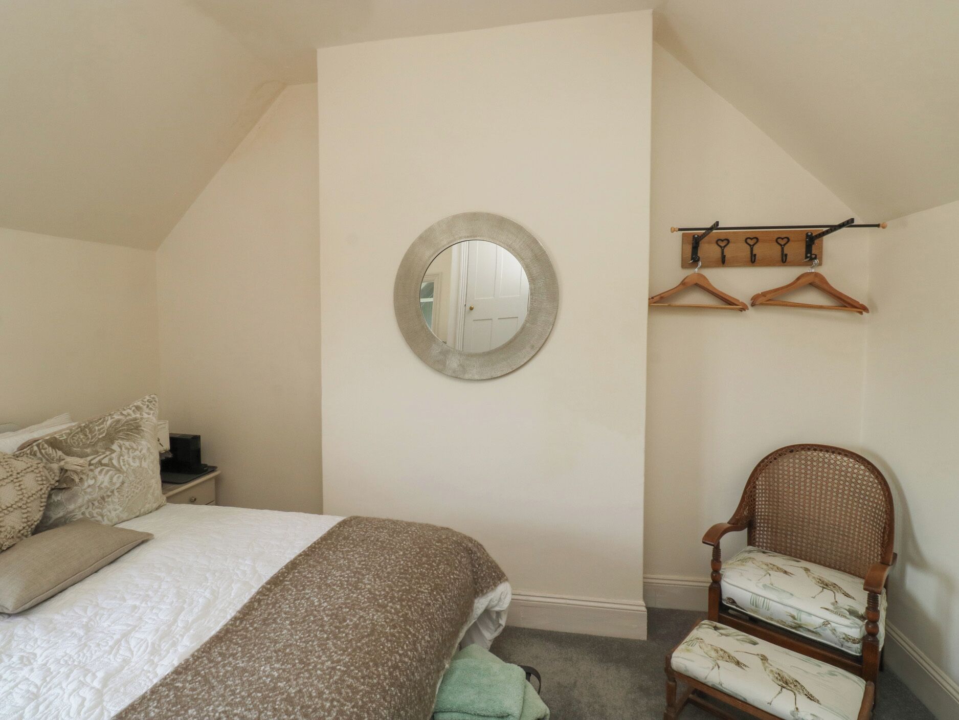 A bedroom with a bed, a round mirror, a chair with a patterned cushion, and a wooden coat rack.