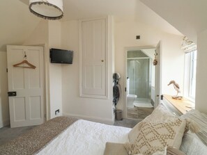 A bedroom with a bed, a TV, a door, a shower, and a window with a decorative item. - Tracey Cottage (Newton Abbot)