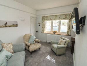 A living room with two armchairs, a sofa, a television mounted on the wall, and a window with floral curtains. - Tracey Cottage (Newton Abbot)