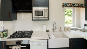 A kitchen with a gas stove, microwave, dishwasher, and a window with a floral curtain. - Tracey Cottage (Newton Abbot)