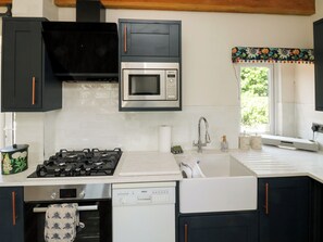 A kitchen with a gas stove, microwave, dishwasher, and a window with a floral curtain. - Tracey Cottage (Newton Abbot)