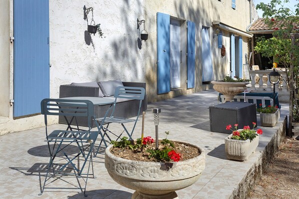 An outdoor seating area with tables and chairs, a large planter with flowers, and a building with blue shutters.