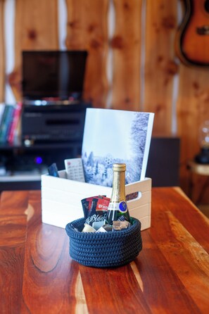 A bottle of champagne in a blue basket on a wooden table with a white box containing a remote control and a book. - Cozy 1-bedroom apartment in peaceful Pohorje with forest and city view (Maribor)